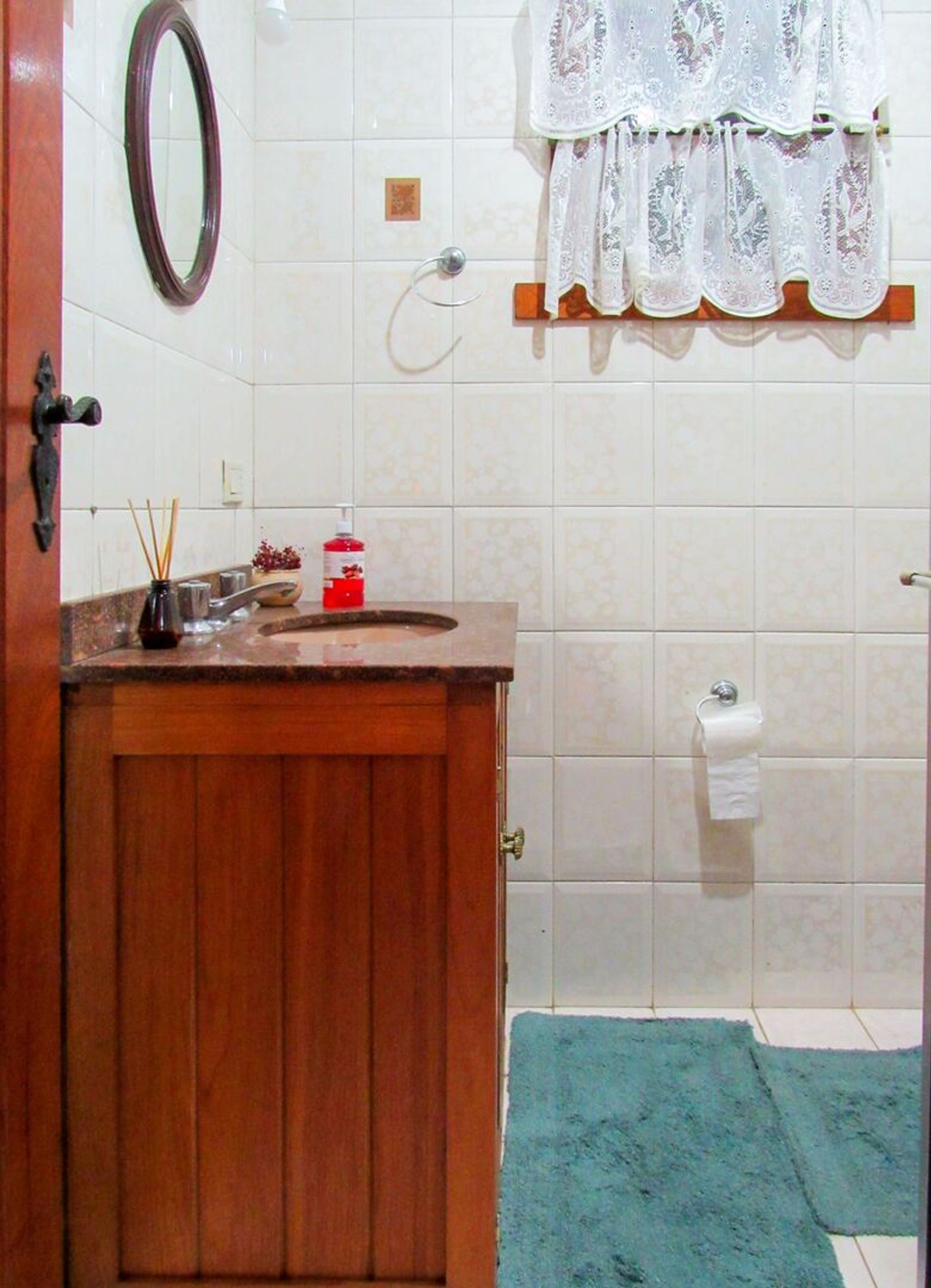 Een badkamer met een houten wastafelmeubel, een spiegel, een handdoekrek met een kantgordijn, een toiletpapierhouder en een blauwe badmat.