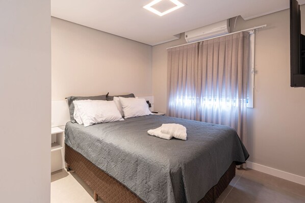 A neatly made bed with a grey quilt, a white pillow, and a white towel on top. There is a white nightstand beside the bed.