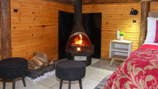 A cozy log cabin room with a central wood-burning stove, a bed with a red and white patterned blanket, and a small white nightstand with a lamp. - Chal com Wi-fi e Muito Lazer em Bom Retiro SC (Bom Retiro)