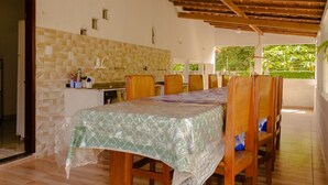 A dining area with a table covered in a patterned tablecloth, wooden chairs, a kitchen with a tiled backsplash, and a wooden ceiling.