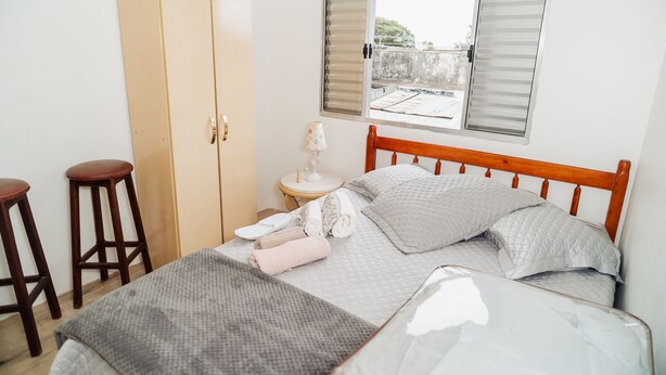 A bedroom with a bed, two wooden stools, a wardrobe, a bedside table with a lamp, and a window with blinds. - Timo Apto c Wifi Ar-condicionado e Garagem (Uruguaiana)