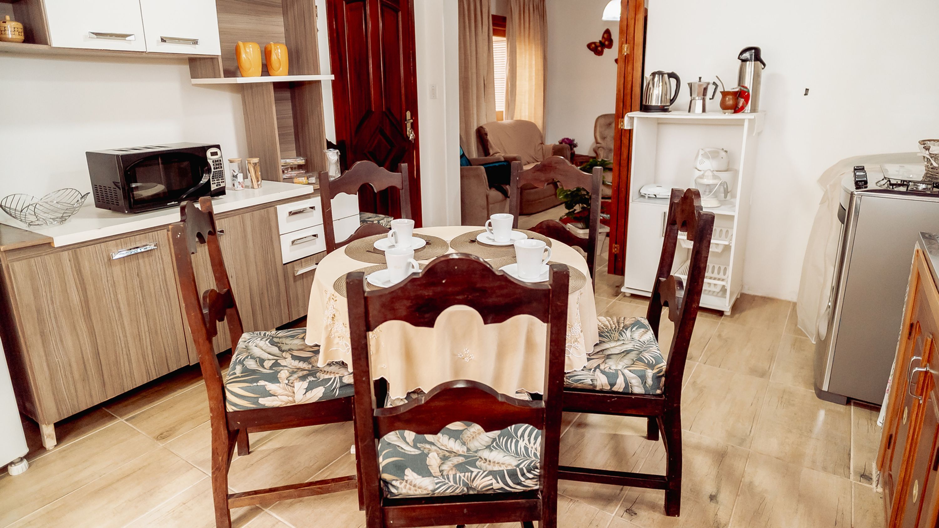 A compact living space with a dining area featuring a round table and four chairs with floral upholstery. A microwave and a small refrigerator are visible in the kitchen area.