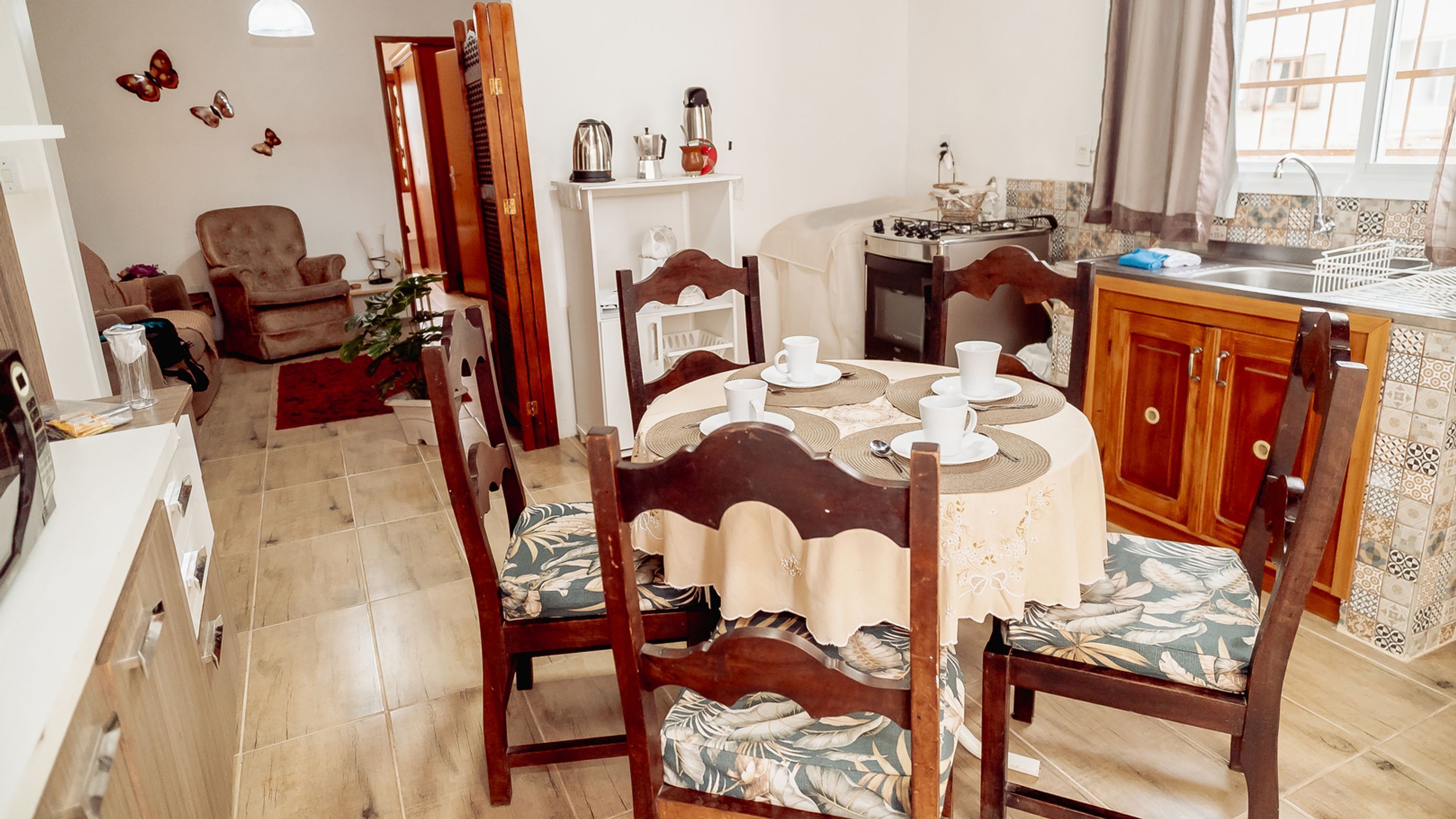 Un comedor con una mesa redonda dispuesta para cuatro personas, rodeada de sillas de madera con tapicería floral. Se observa una cocina con estufa a gas y diversos electrodomésticos al fondo.