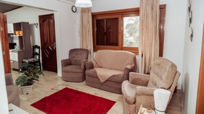 A living room with a red rug, two brown armchairs, a sofa, a dining area with a kitchen, and a large window with curtains.