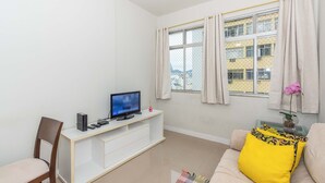 A living room with a TV on a white stand, a chair, a sofa with a yellow pillow, and a window with curtains. - Apto com Wifi e Tima Localiza o Laranjeiras RJ (Rio de Janeiro)