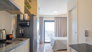 Ein kompaktes Hotelzimmer mit Küchenzeile, einem Bett, einem kleinen Tisch und einem Balkon mit Vorhängen.