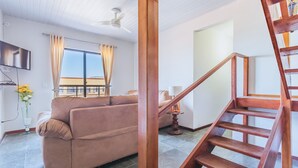 Un salon comprenant un canapé, un téléviseur fixé au mur, un ventilateur de plafond et un escalier avec des rampes en bois.