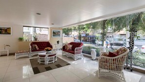 A spacious room with white wicker furniture, a red sofa, and a view of the street through large windows. - Apto Top Pitangueiras no Melhor Ponto do Guaruj (Guarujá)