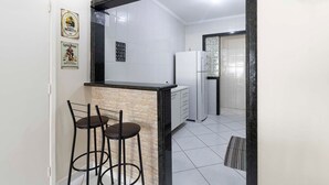A kitchen with a marble countertop, two bar stools, a refrigerator, and a tiled backsplash. - Apto Top Pitangueiras no Melhor Ponto do Guaruj (Guarujá)