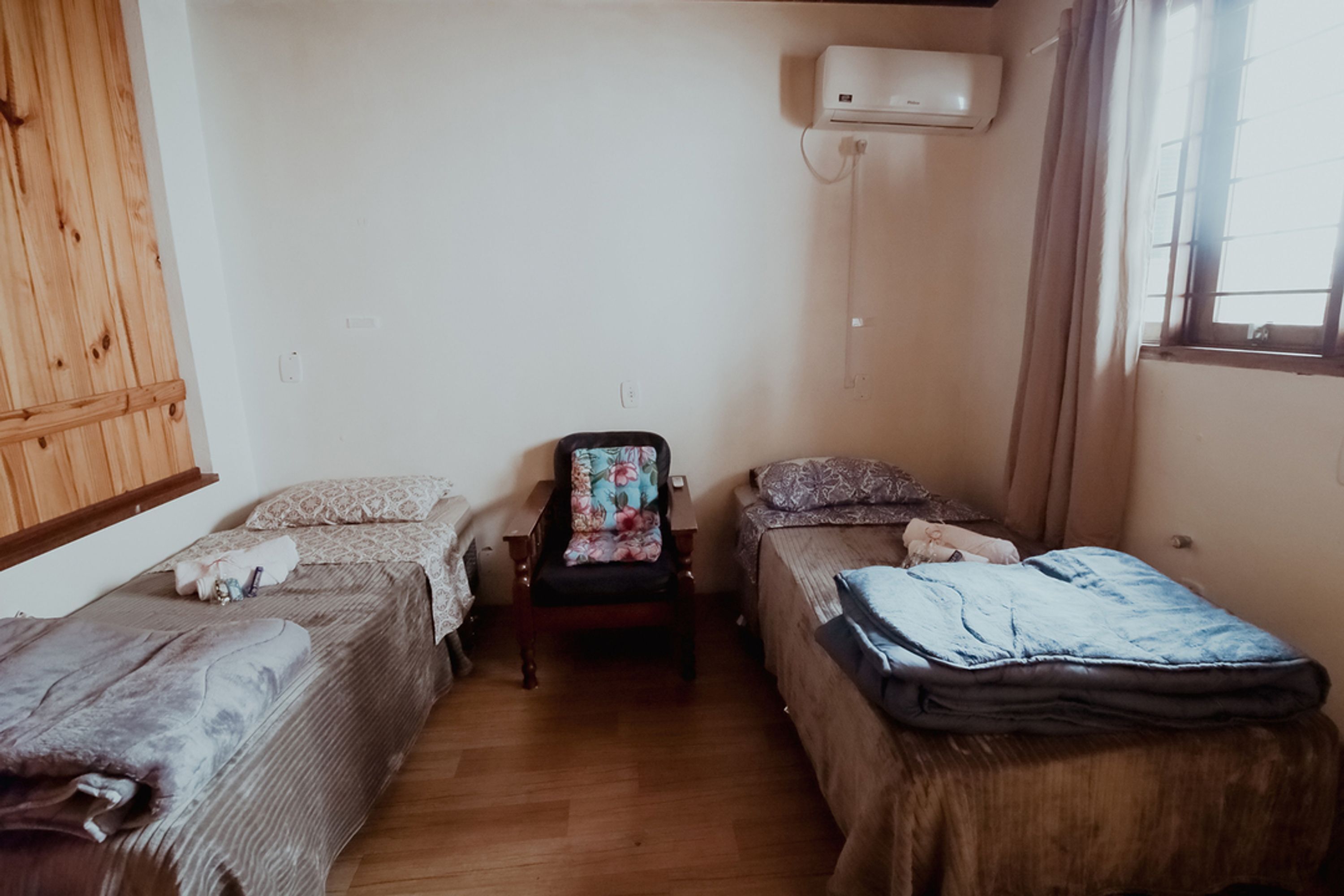 Una habitación pequeña y sencilla con dos camas individuales, una silla de madera, un equipo de aire acondicionado y una ventana con cortinas.