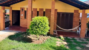 Edificio a un piano con facciata gialla, zoccolo in mattoni e tetto in tegole. Nel giardino anteriore si nota un piccolo arbusto verde e un'amaca tesa tra due colonne.