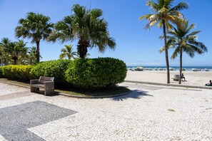 A paved walkway with a bench and palm trees, leading to a beach with umbrellas and people. - Apto Confort vel a 170m da Praia Vila Tupi (Praia Grande)