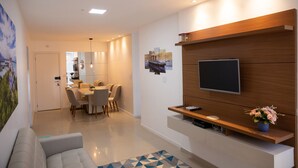 Un soggiorno moderno con una TV a schermo piatto montata su un pannello di legno, un'area pranzo con tavolo e sedie, e un angolo salotto accogliente con divano.
