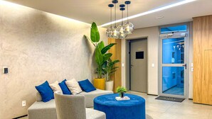 A modern reception area with a grey sofa, blue cushions, a round blue ottoman, and a potted plant. - Excelente Apto a 50m do Metr Butant em S o Paulo (São Paulo)