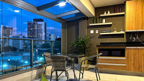 A modern kitchen with a view of the city at dusk, featuring a dining area with chairs and a table, and a wooden cabinet with a sign above it. - Excelente Apto a 50m do Metr Butant em S o Paulo (São Paulo)