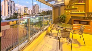 A balcony with a glass railing, a table, and chairs, overlooking a cityscape with tall buildings. - Excelente Apto a 50m do Metr Butant em S o Paulo (São Paulo)