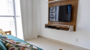 A modern living room with a flat-screen TV mounted on a wooden wall, a sofa with a floral-patterned throw, and a window with curtains. - Apto Confort vel a 150m do mar Completo com Lazer (Praia Grande)