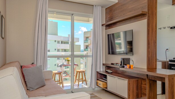 A modern living room with a sofa, a TV mounted on the wall, a wooden cabinet, and a balcony with a view of buildings. - Apartamento Completo em Trindade Perto da Ufsc (Florianópolis)