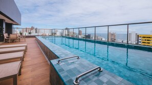 Piscina infinita no terraço com vista para a cidade, deck de madeira e espreguiçadeiras.