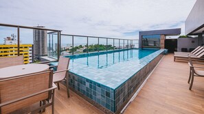 Uma piscina infinita no terraço, com borda de vidro, rodeada por espreguiçadeiras de madeira e um espaço para refeições.