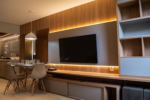 A modern living room with a flat-screen TV mounted on a wooden wall, a dining area with a table and chairs, and built-in shelves. - Apto c Wi-fi Tima loc Prox ao Eixo Monumental (Brasília)