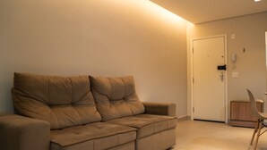A modern living room with a beige sofa, a wooden side table, and a door with a phone. - Apto c Wi-fi Tima loc Prox ao Eixo Monumental (Brasília)