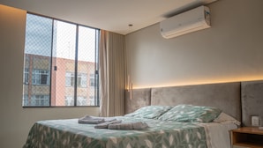 A bedroom with a bed, a nightstand, a window with curtains, and an air conditioning unit. - Apto c Wi-fi Tima loc Prox ao Eixo Monumental (Brasília)