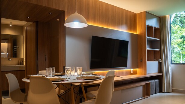 A modern dining area with a wooden table, white chairs, a flat-screen TV, and a pendant light. - Apto c Wi-fi Tima loc Prox ao Eixo Monumental (Brasília)