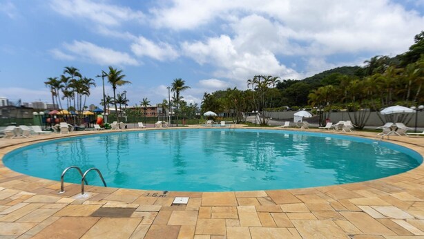 A large, oval-shaped swimming pool with a tiled deck, surrounded by palm trees and lounge chairs. - Casa em Condom nio com Piscina Wi-fi e Garagem (Guarujá)