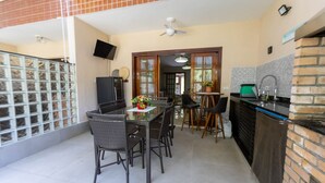 A dining area with a glass table and wicker chairs, a kitchen with a brick backsplash, and a ceiling fan. - Casa em Condom nio com Piscina Wi-fi e Garagem (Guarujá)