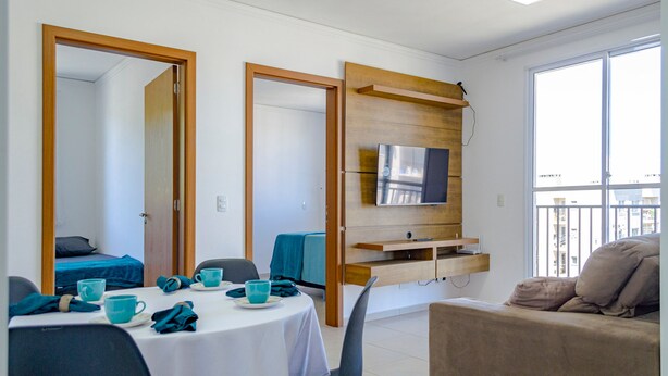 A modern living room with a dining area, a sofa, a TV mounted on a wooden panel, and a balcony with a railing. - Apto c Tima Localiza o Prox Praias e pq Aquatico (Florianópolis)