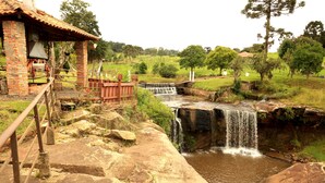 Un pont de pierre rustique enjambant une petite cascade, un bâtiment en briques doté d’un balcon et un paysage verdoyant luxuriant.