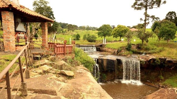 Un pont de pierre rustique enjambant une petite cascade, un bâtiment en briques doté d’un balcon et un paysage verdoyant luxuriant.