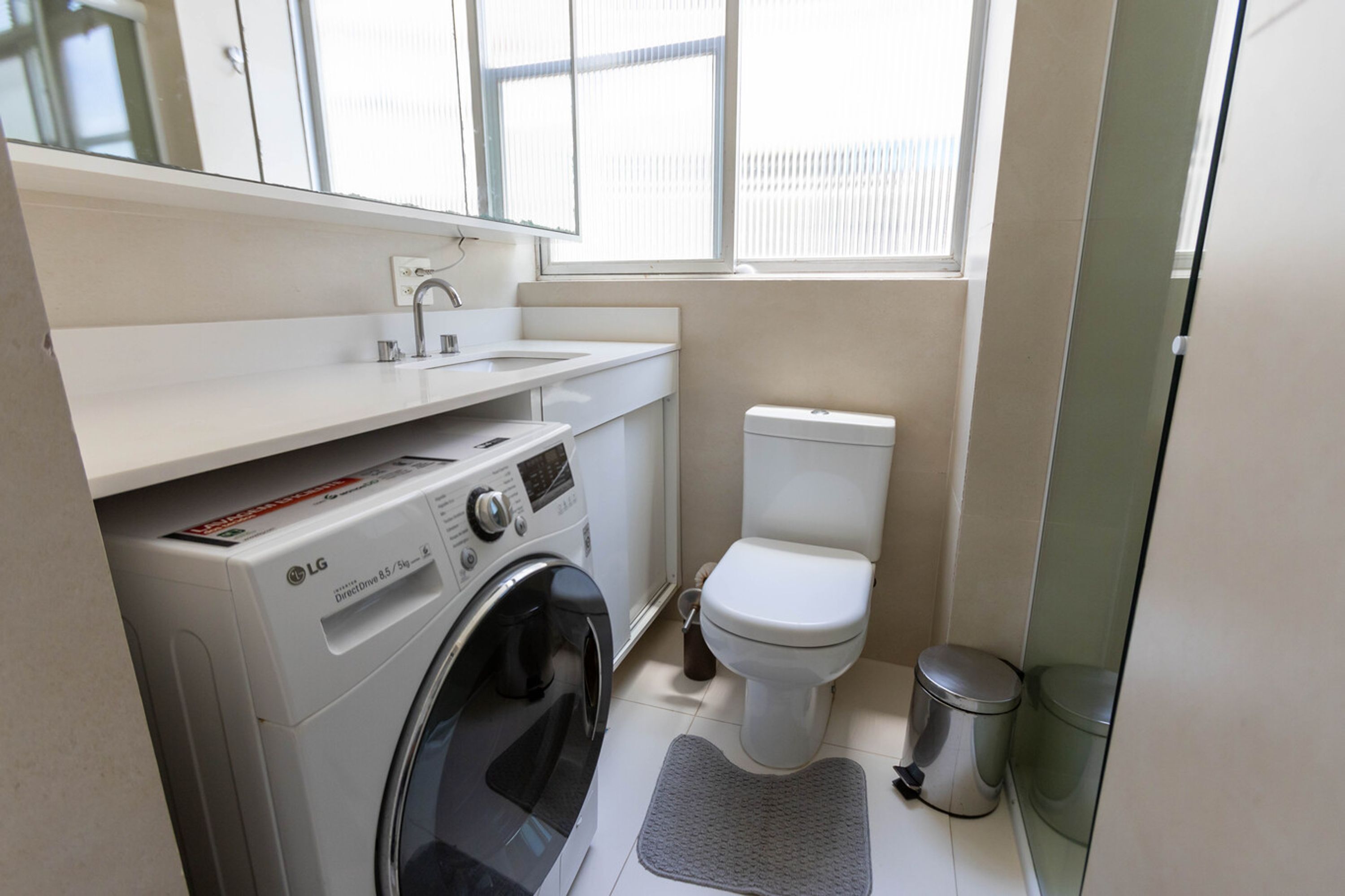 A compact laundry room with a washing machine, a sink, a toilet, and a trash bin.