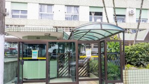 A glass-enclosed entrance with a metal roof, a tiled floor, and a staircase leading to a building with a green awning. - Apto Confort vel com Linda Vista Frente Para o mar (Santos)