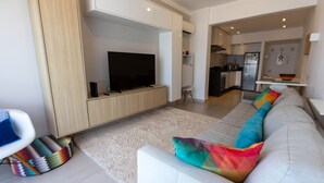 A modern living room with a flat-screen TV, a white sofa with colorful cushions, and a kitchen in the background. - Apto Confort vel com Linda Vista Frente Para o mar (Santos)