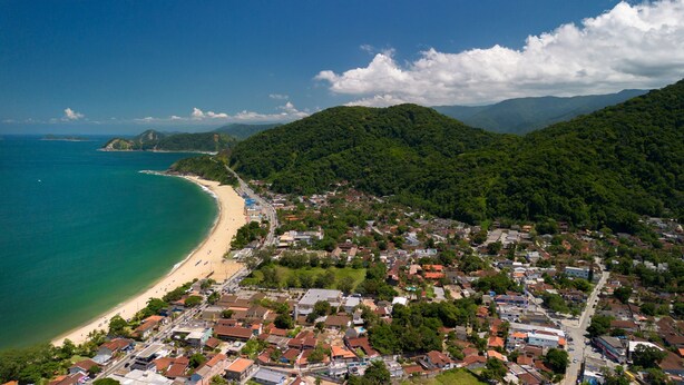 Vista aérea de un pueblo costero con playa, zonas residenciales y colinas boscosas.