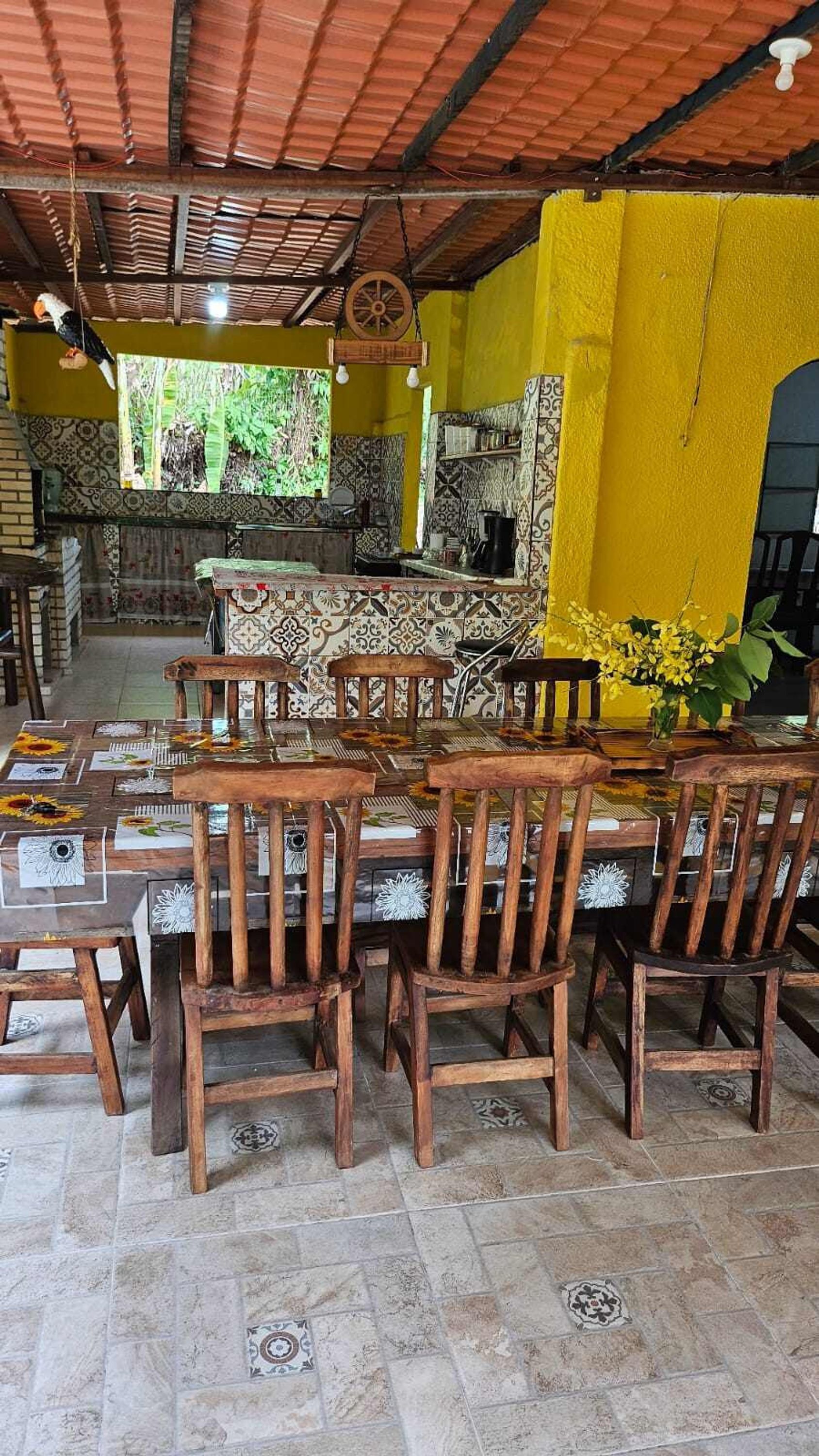 Uma área de refeições com uma mesa de madeira comprida e cadeiras, cercada por paredes amarelas e ladrilhos estampados.