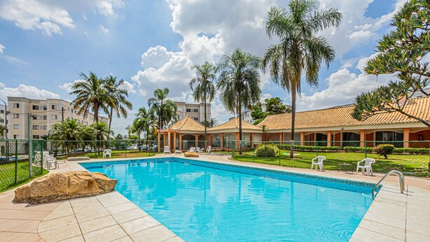 A swimming pool with a tiled deck, surrounded by palm trees and a building with a tiled roof. - Confort vel Apartamento com Wi-fi pet Friendly (Sorocaba)