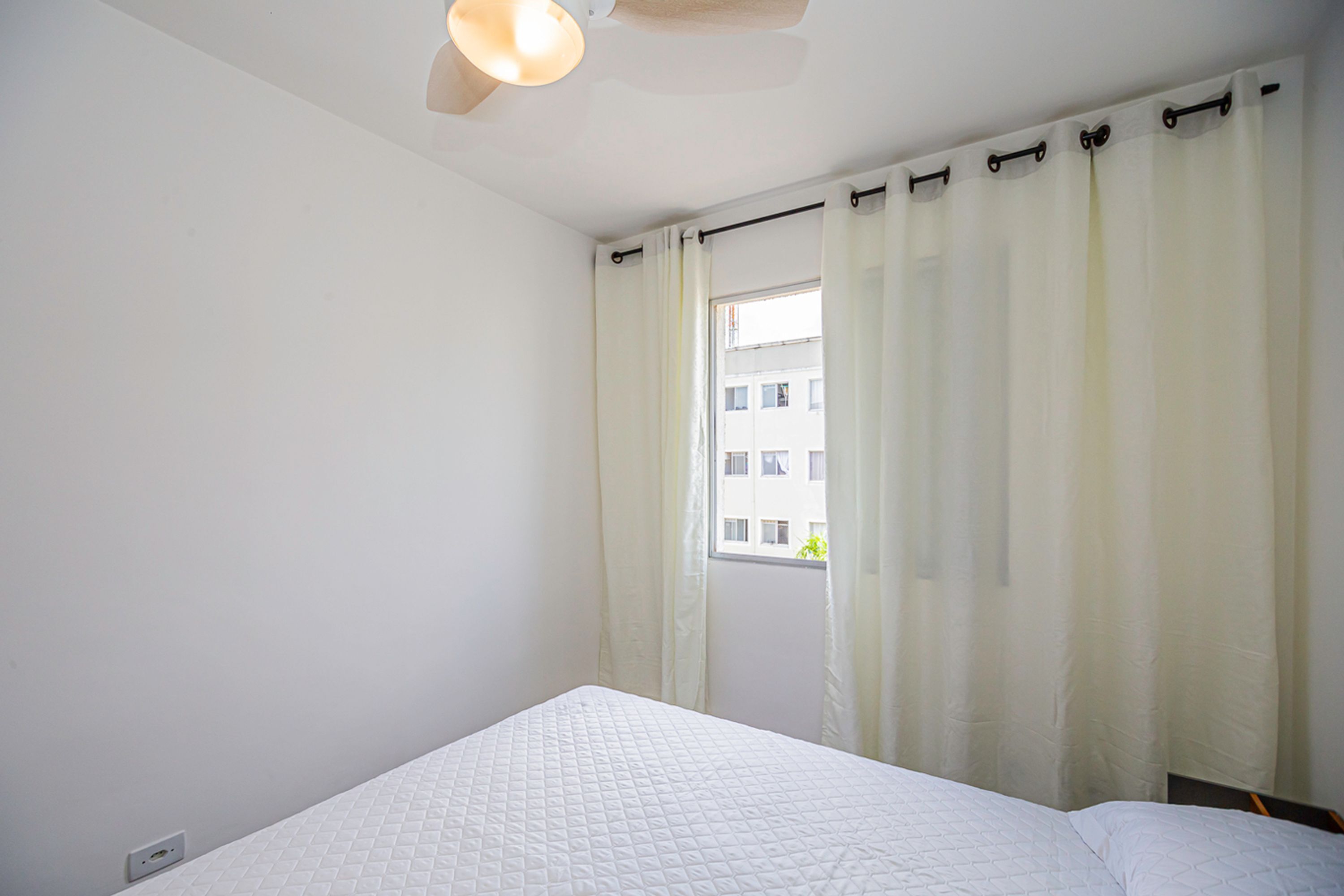 Een eenvoudige, schone kamer met een bed, een raam met gordijnen en een plafondventilator.