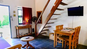 Une pièce comprenant une table à manger, des chaises, une petite table ronde, un escalier en bois, un téléviseur fixé au mur et une fenêtre avec des rideaux.