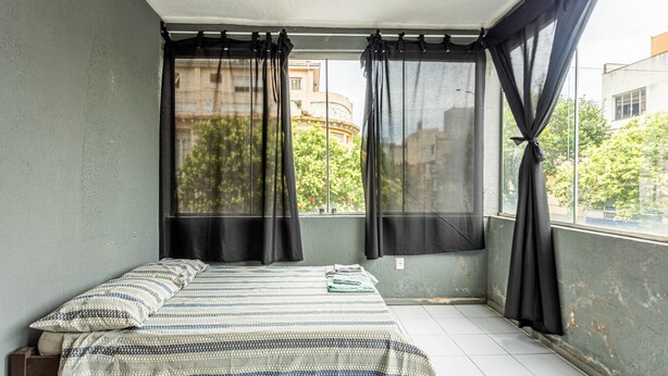 A small, clean room with a bed, a window with black curtains, and a view of buildings and trees outside. - Casa Triplex com Tima Localiza o Pet Friendly (Porto Alegre)