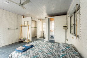 Une pièce aux murs en briques peintes en blanc, un lit avec une literie à motifs, un ventilateur de plafond et une fenêtre avec des stores.