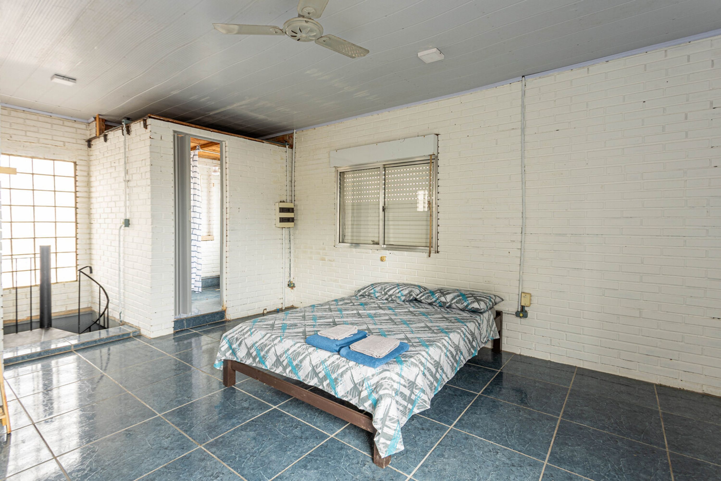 A bedroom with a bed, a ceiling fan, a window with blinds, and a door leading to another room.