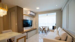 A modern living room with a flat-screen TV, a dining area with a white table and chairs, and a large window providing natural light. - Apto Confort vel com Wi-fi e Churrasqueira (Fortaleza)