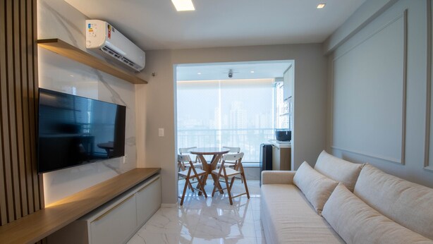 A modern living room with a flat-screen TV, a dining table with chairs, a sofa, and a large window offering a city view. - Apto Confort vel com Wi-fi e Churrasqueira (Fortaleza)