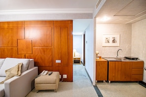 A hotel room with a wooden wardrobe, a sofa, an ottoman, a small bedside table with a lamp, and a framed picture on the wall.