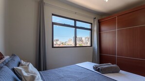 A hotel room with a large bed, a wooden wardrobe, a window with a city view, and a curtain. - Apto Confort vel a 450m do mar da Praia do Morro (Guarapari)