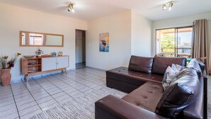 A modern living room with a brown leather sofa, a wooden cabinet, and a large window with a view of buildings. - Apto Confort vel a 450m do mar da Praia do Morro (Guarapari)