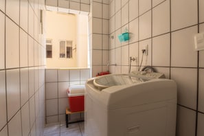 A laundry room with a washing machine, a red cooler, and a window providing natural light. - Apto Confort vel a 450m do mar da Praia do Morro (Guarapari)